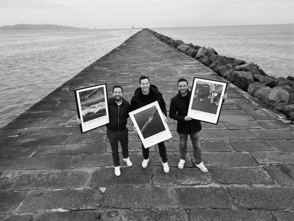 Black and white photo of the three founding members of Love Your Spot standing on a pier each holding one of their framed prints
